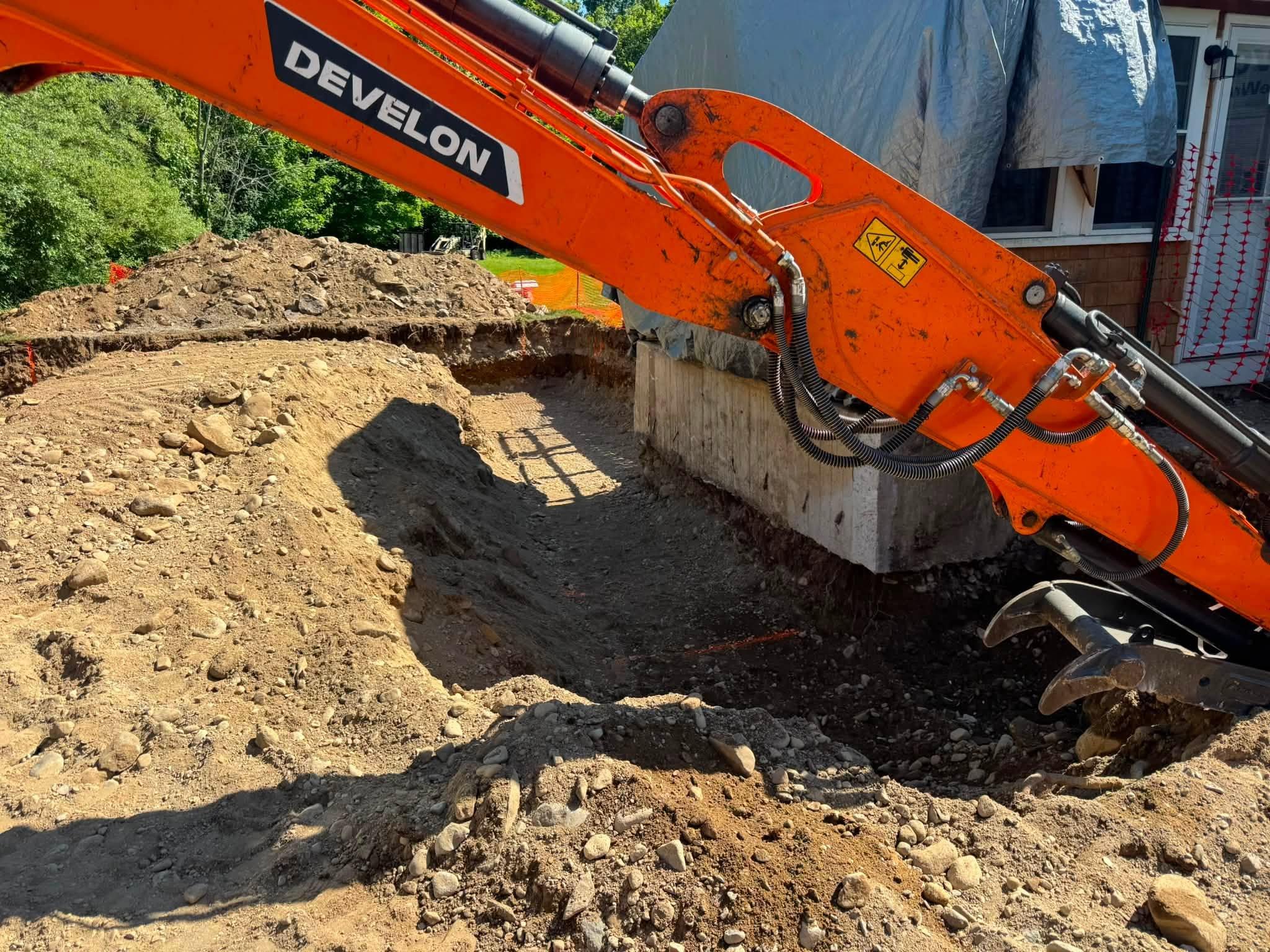 Orange excavator digging immediately beside an existing residential foundation