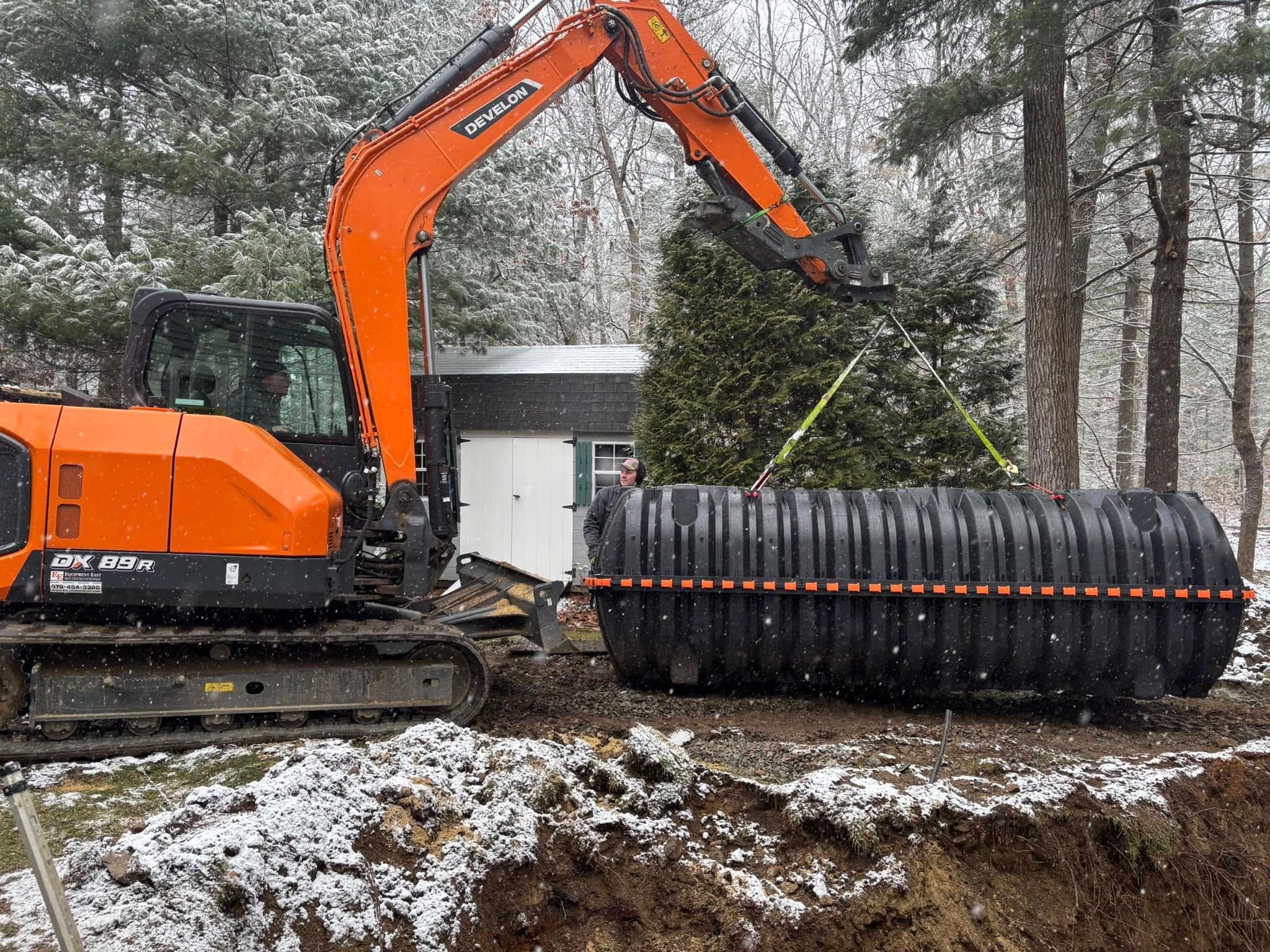 Excavator lifting a large tank beside a residential work area in falling snow